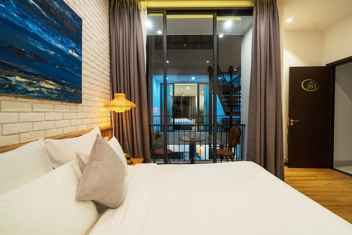 A hotel room with a bed, a painting on the wall, a balcony with a table and chairs, and a door with a number.