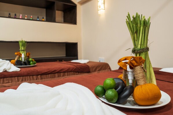 Habitación de hotel con dos camas, una mesita de noche con una bandeja de frutas y una botella de loción, y un estante en la pared con objetos decorativos.