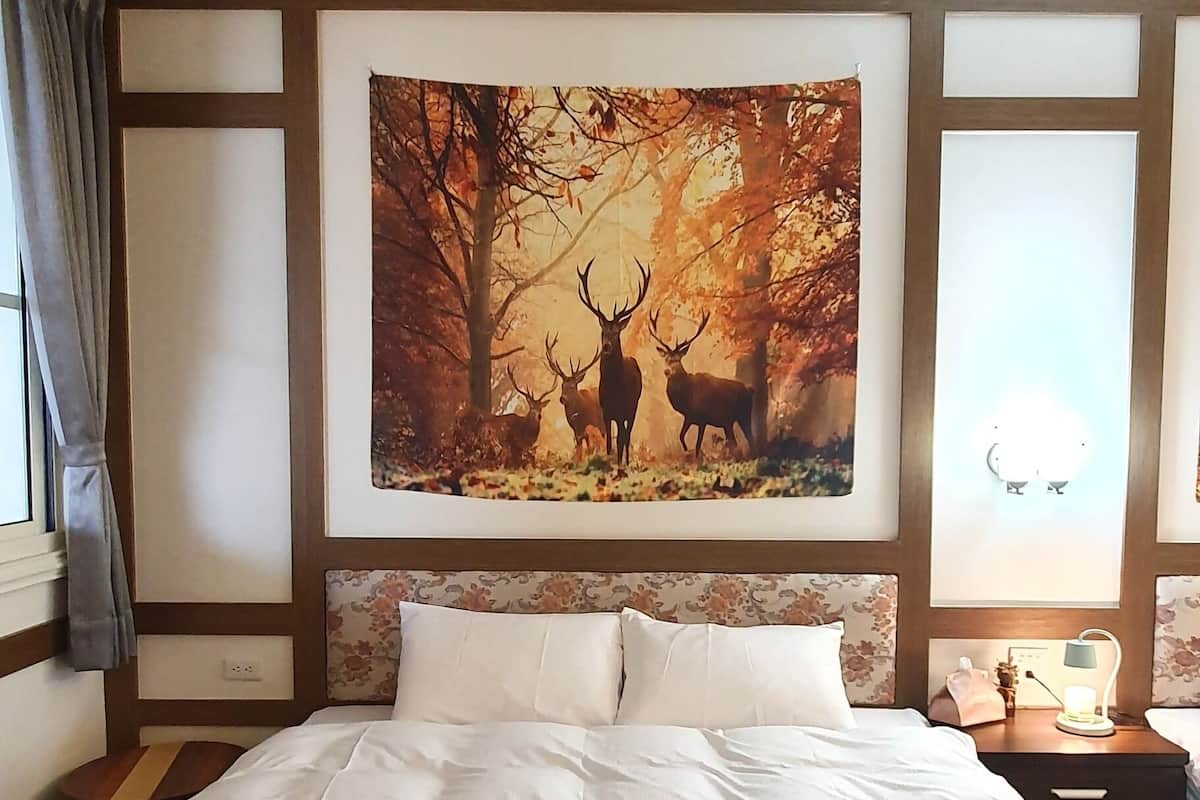 Una cama grande con sábanas blancas, un cuadro de ciervos en follaje otoñal en la pared, una mesita de noche con una lámpara y una ventana con cortinas.