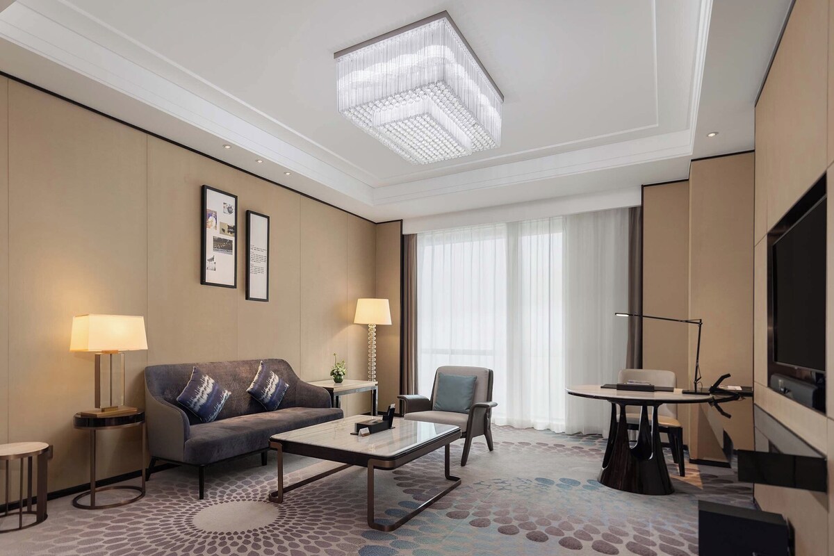 A modern hotel room with a sofa, coffee table, armchair, and a TV.