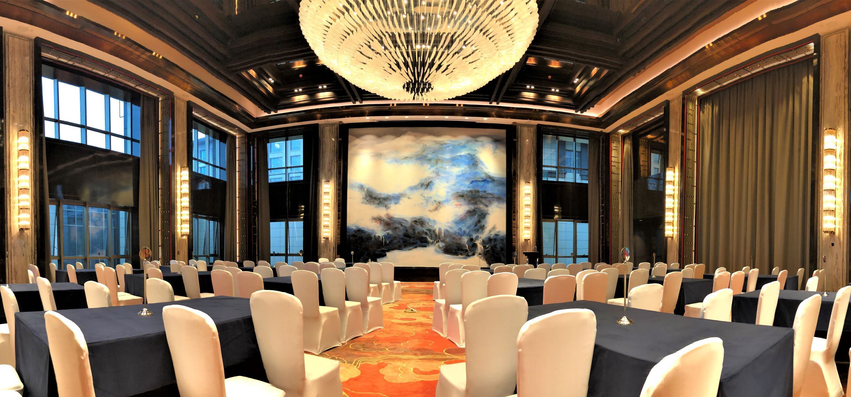 A conference room with a large screen, rows of tables, and chairs, featuring a chandelier and large windows.