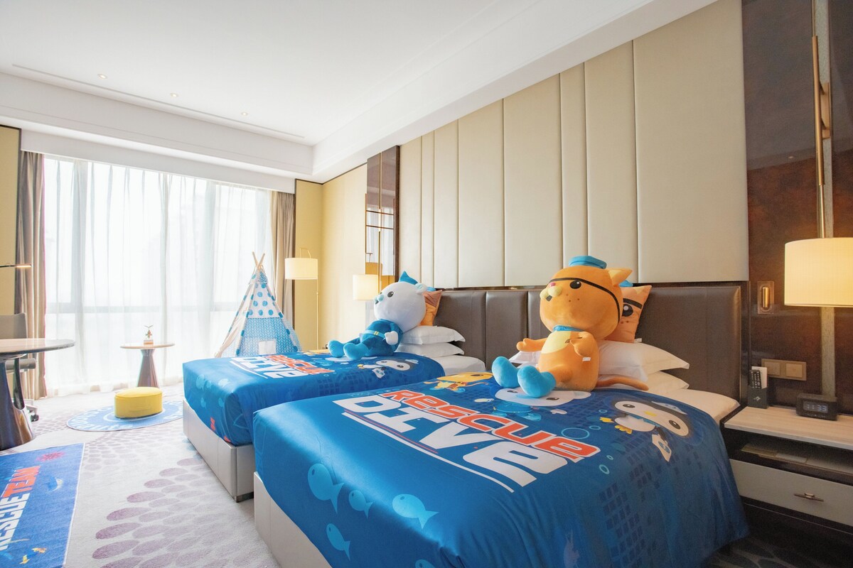A hotel room with two beds, each with a blue bedspread featuring a cartoon character. There is a small table with a lamp, a window with curtains, and a nightstand with a lamp.