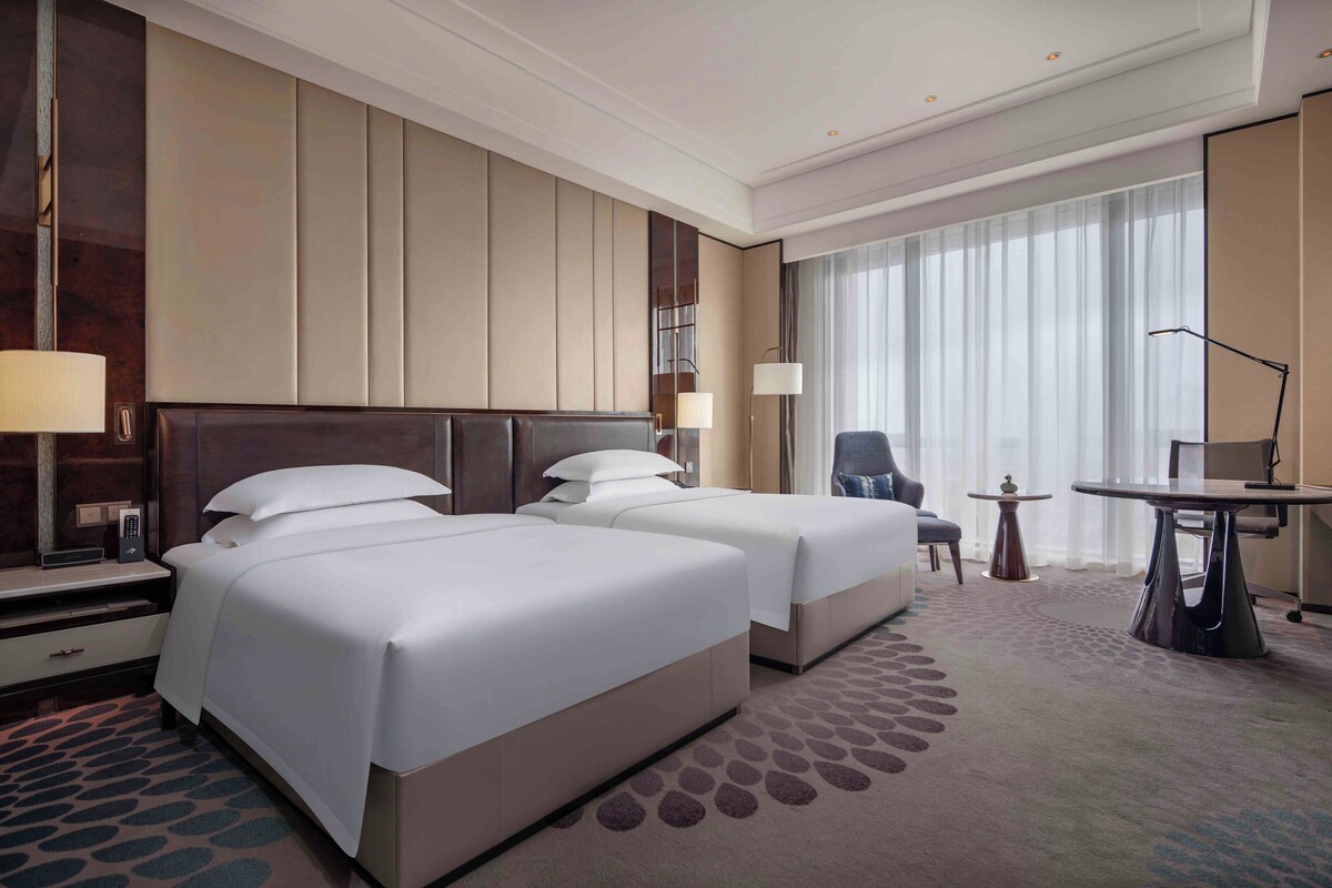 A hotel room with two beds, a desk, a chair, and a large window with curtains.