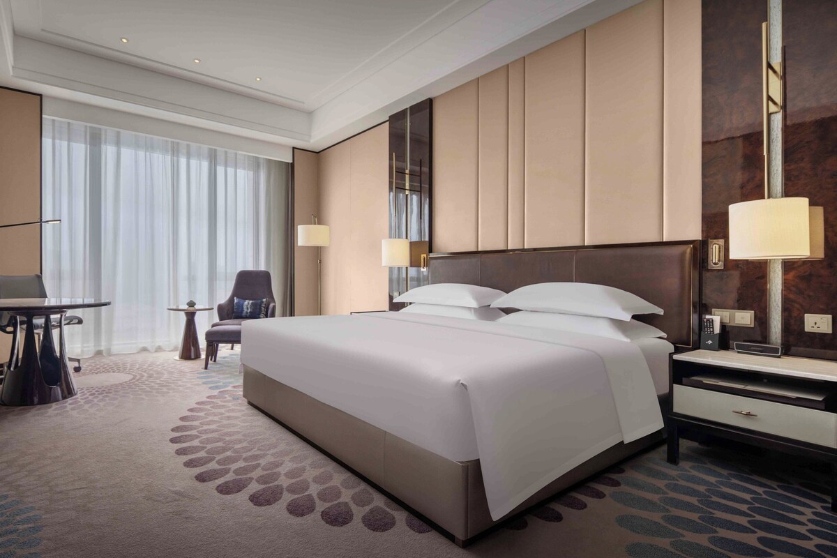 A hotel room with a large bed, a desk, a chair, and a nightstand with a lamp.