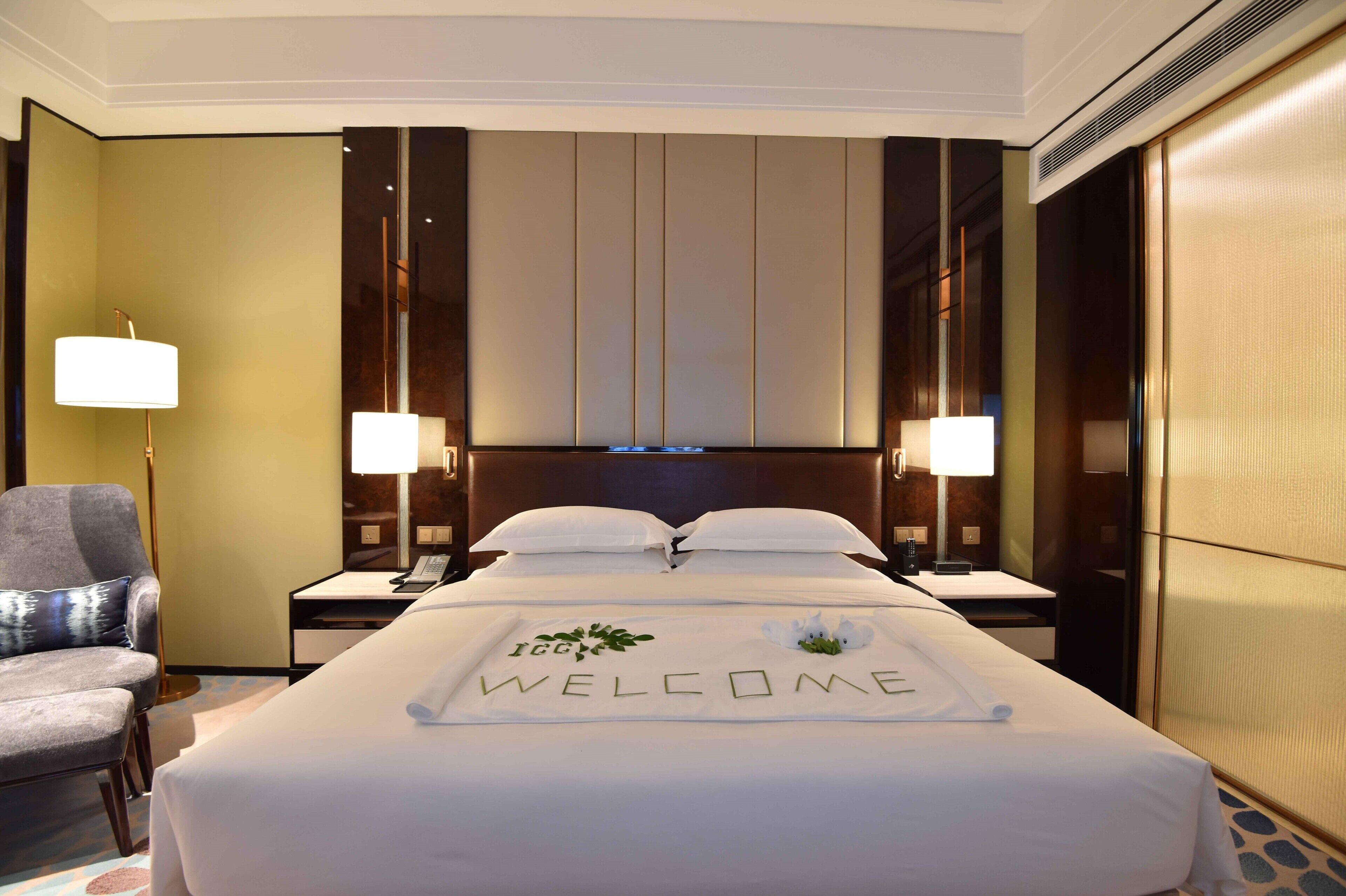 A hotel room with a large bed, two bedside tables, a chair, and a nightstand. The bed has a welcome mat with a floral design.