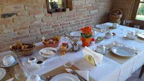 A dining table set with white plates, bowls, and cutlery, a basket of bread, a vase with flowers, and a window with a view of greenery. - Agriturismo il Querciolo (Marciano della Chiana)