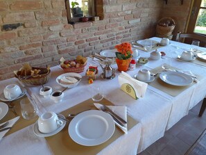 A dining table set with white plates, bowls, and cutlery, a basket of bread, a vase with flowers, and a window with a view of greenery. - Agriturismo il Querciolo (Marciano della Chiana)
