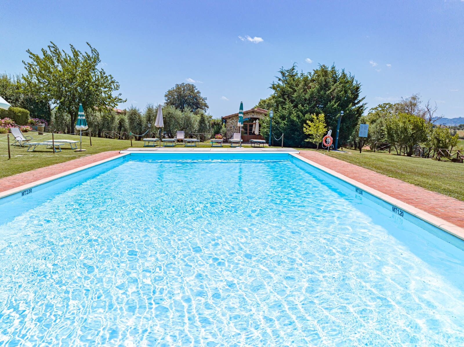 Una piscina rectangular con agua azul cristalina, rodeada por un camino de ladrillo y césped verde. Hay sillas de descanso y sombrillas a lo largo de la piscina.