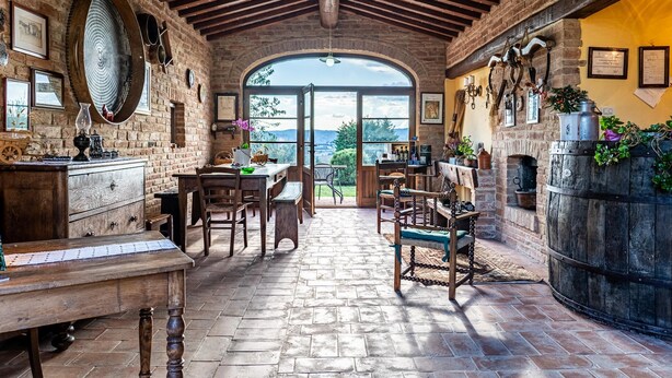Ein rustikales Interieur mit Kamin aus Stein, Holzmöbeln und einem großen Bogenfenster mit Blick ins Freie.