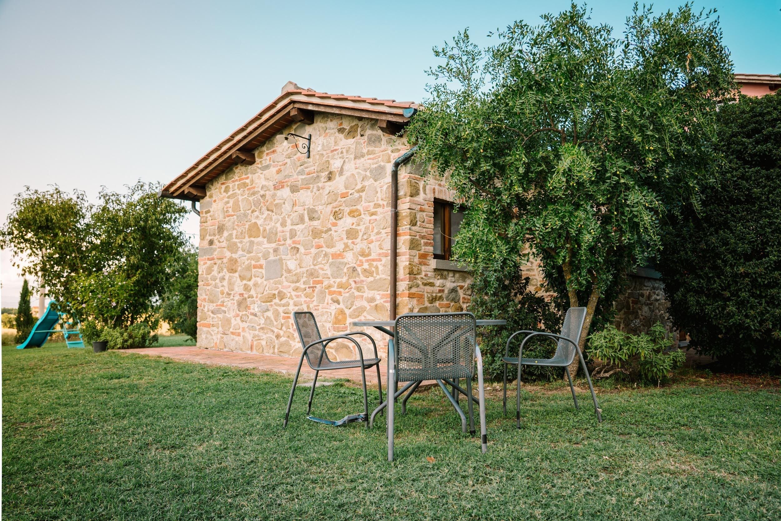 A stone building with a tiled roof, surrounded by a grassy area with metal chairs and a small table.