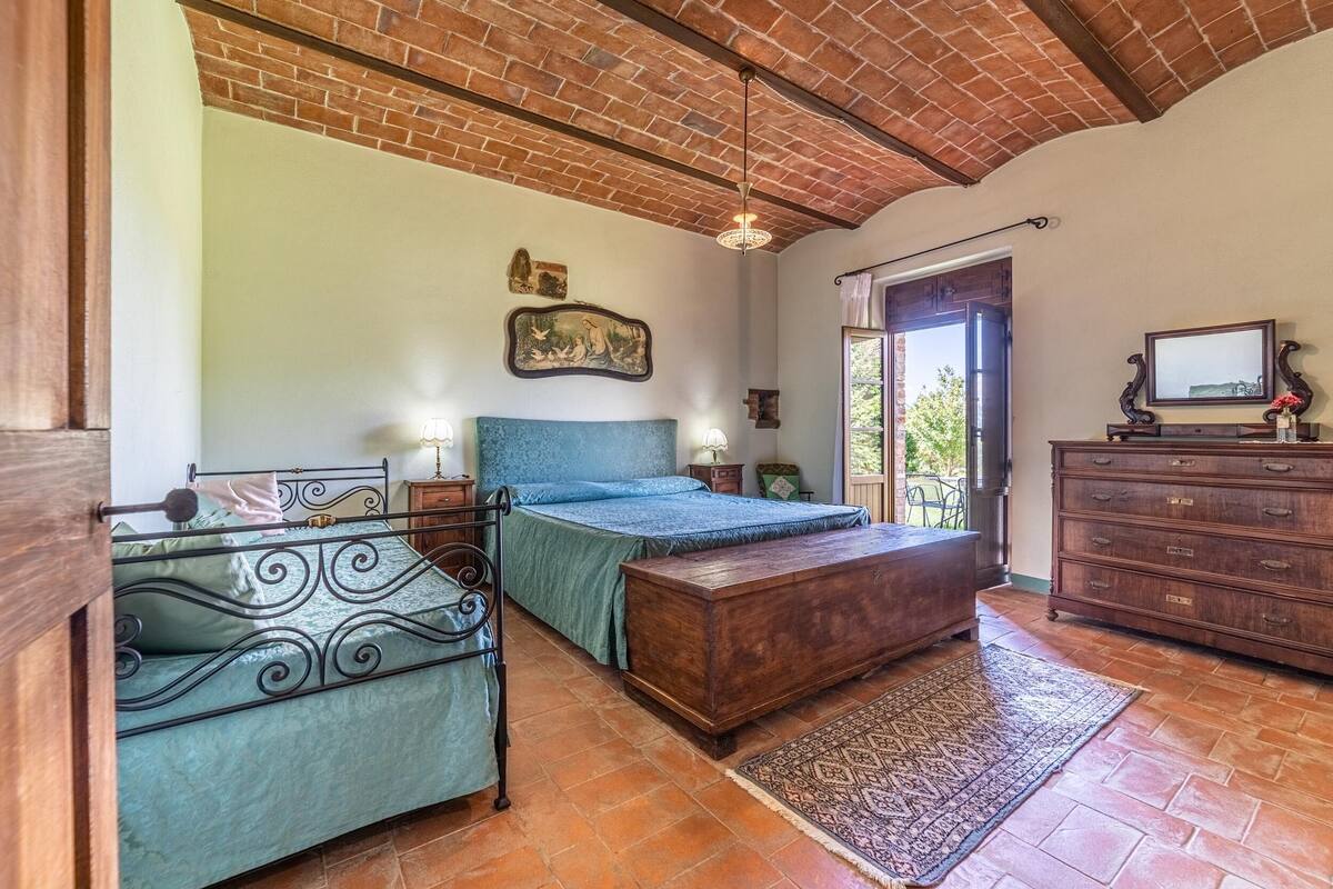 A bedroom with a brick ceiling, a bed with a blue headboard, a wooden dresser, and a small television on a dresser.