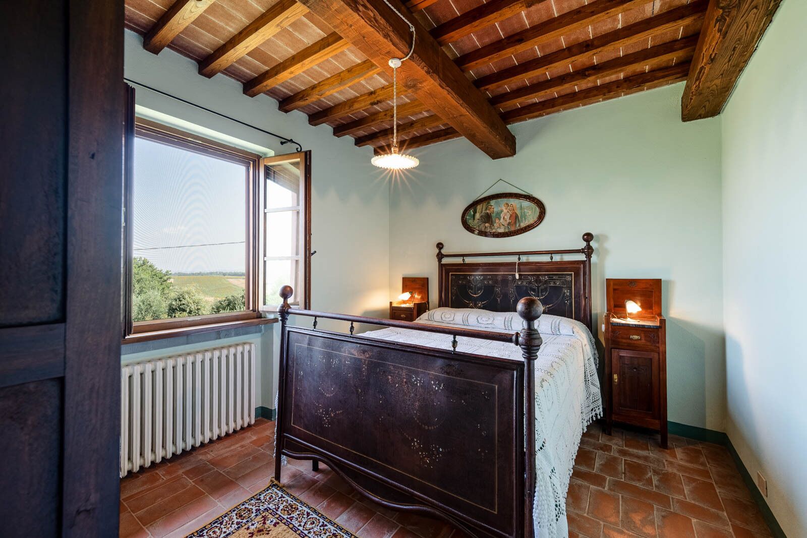 A bedroom with a wooden bed, a radiator, a window with a view, and a hanging light fixture.