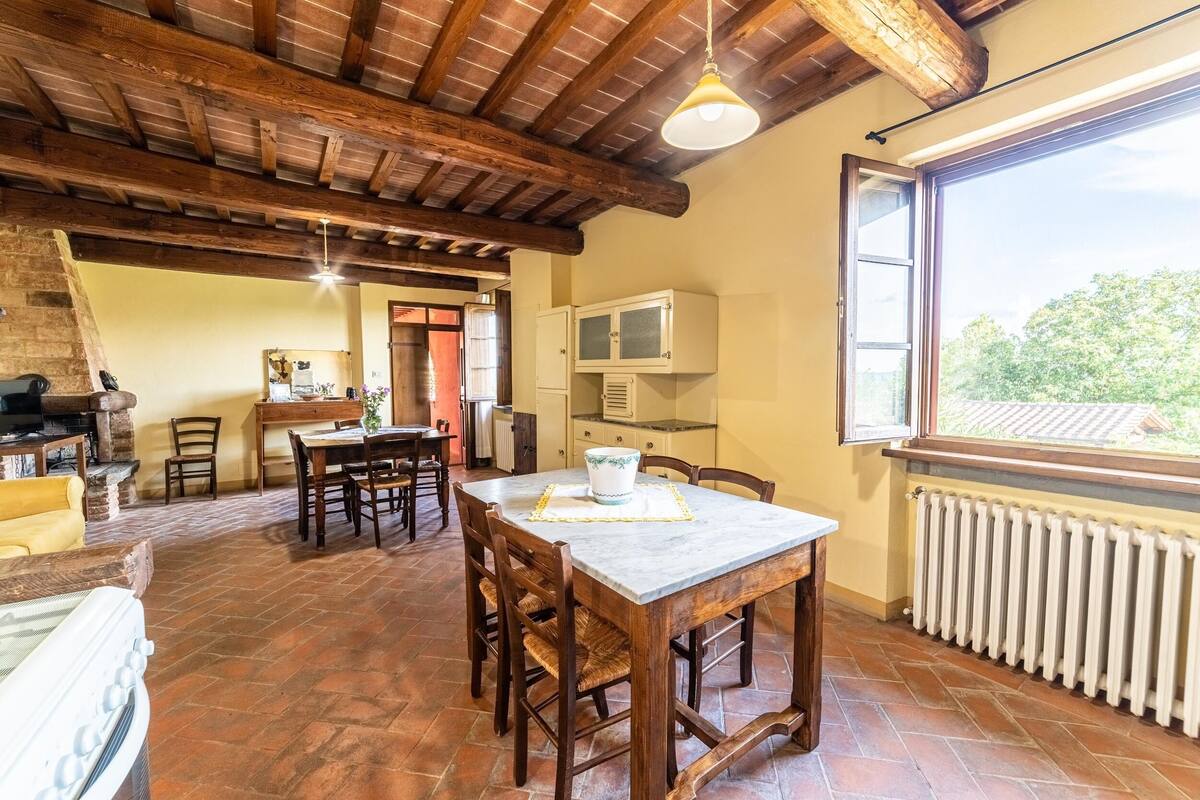 A spacious dining area with a wooden table and chairs, a kitchen area with cabinets, a radiator, and a window with a view of greenery.