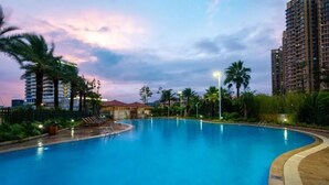 A large swimming pool with a curved edge, surrounded by palm trees and tall buildings. - Yinghai Hotel Apartment Zhuhai Chimelong (Zhuhai)