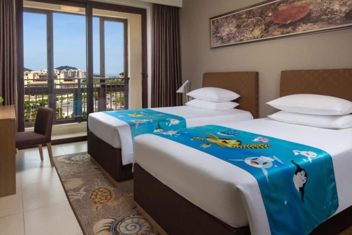 Ein Hotelzimmer mit zwei Betten, einem Schreibtisch und einem Sessel. Ein großes Fenster bietet Blick auf Gebäude und die Stadtlandschaft.