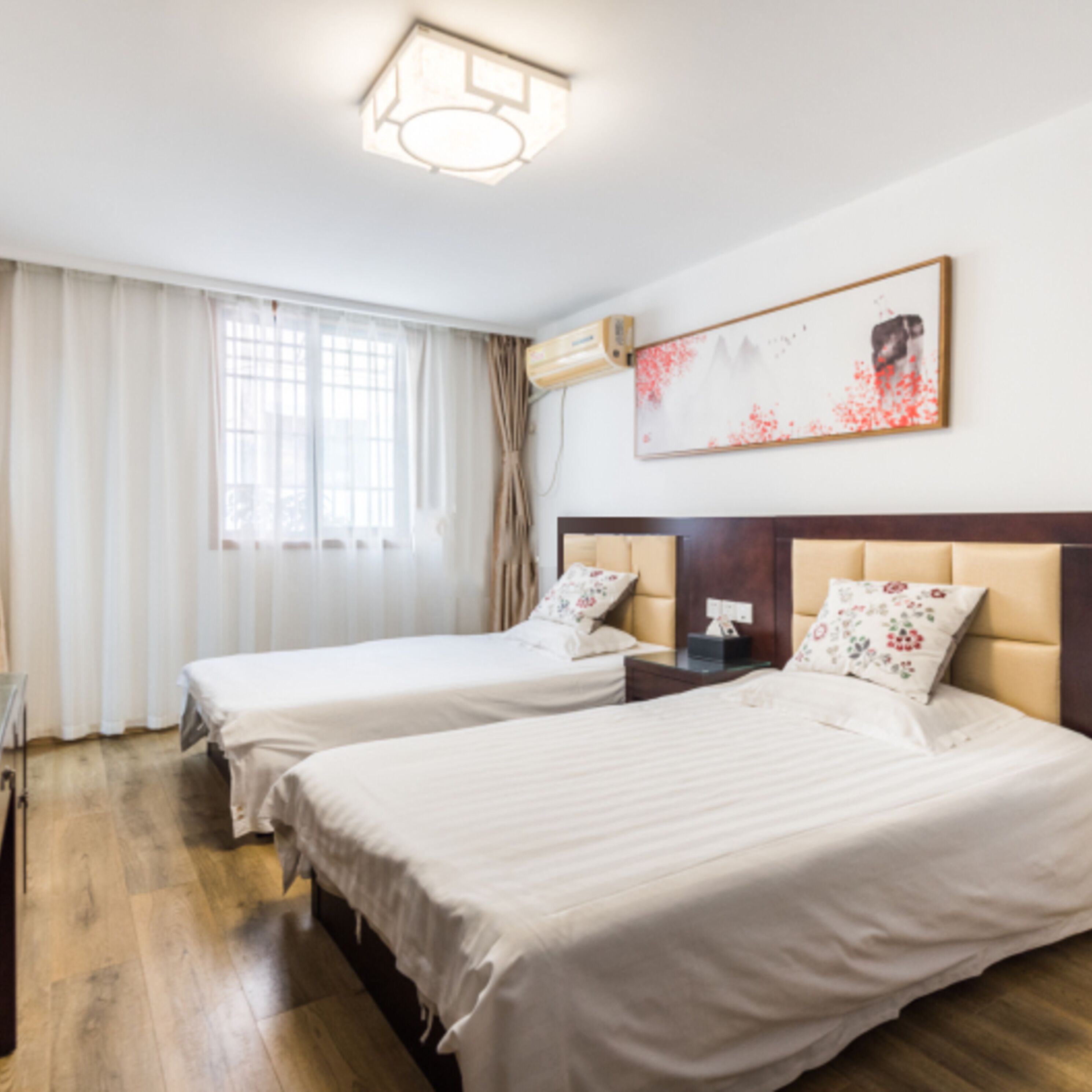 A hotel room with two beds, a window with curtains, a painting on the wall, and an air conditioning unit.