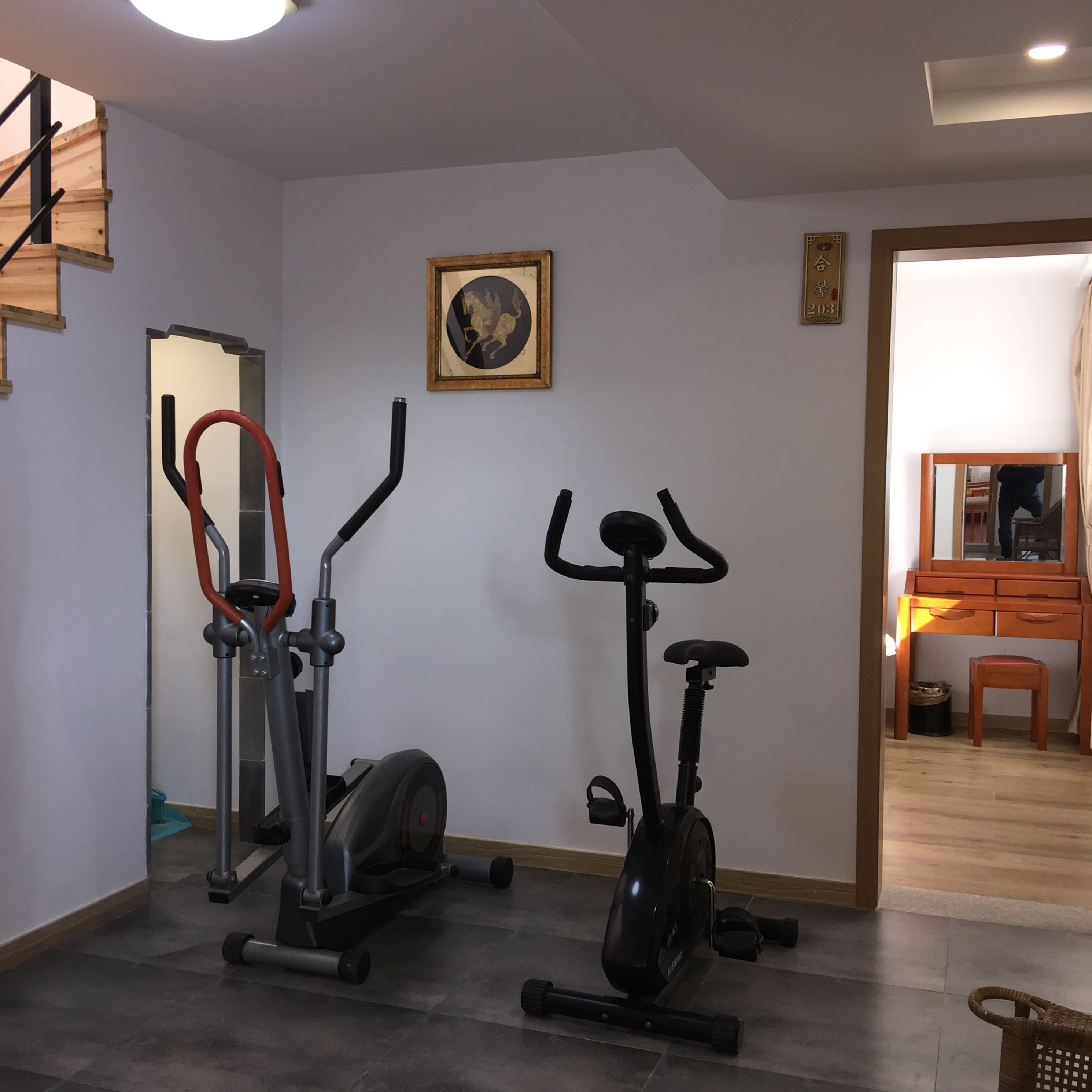 A home gym with two exercise bikes and a stationary bike, a staircase, and a mirror reflecting the room.