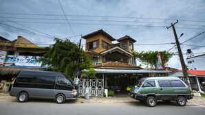 A two-story building with a tiled roof and a balcony. There are two cars parked in front, and a fence with geometric patterns. - Sana's Beach Guesthouse and Restaurant (Negombo)