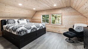 Un dormitorio con techo de madera, una cama con ropa de cama estampada, una silla negra y una ventana con vista a árboles.