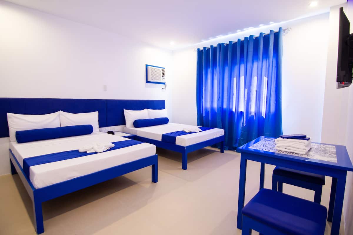 A hotel room with two beds, a small table with a chair, and a window with blue curtains.