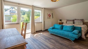 A bedroom with a bed, a blue sofa, a wooden table, and a window with a view of houses and trees. - Old forest house in the Eifel (Ormont)