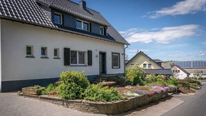 A two-story house with a garden in front, a paved driveway, and a wind turbine in the background. - Old forest house in the Eifel (Ormont)