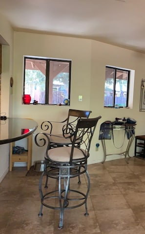 A dining area with a round table and two chairs, a window with a view of the outdoors, and a small shelf with items.
