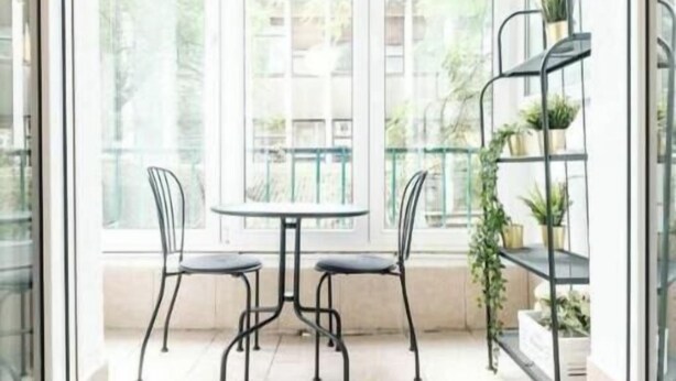 Une terrasse lumineuse et spacieuse, avec une porte vitrée donnant sur un balcon. On y trouve deux chaises en métal noir et une petite table ronde. Un présentoir métallique avec des plantes en pot est visible sur la droite.