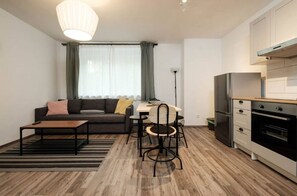 Ein kompakter Wohnbereich mit Sofa, Esstisch, Stühlen, Couchtisch, Küchenzeile mit Kühlschrank und Backofen sowie einem Fenster mit Vorhängen.