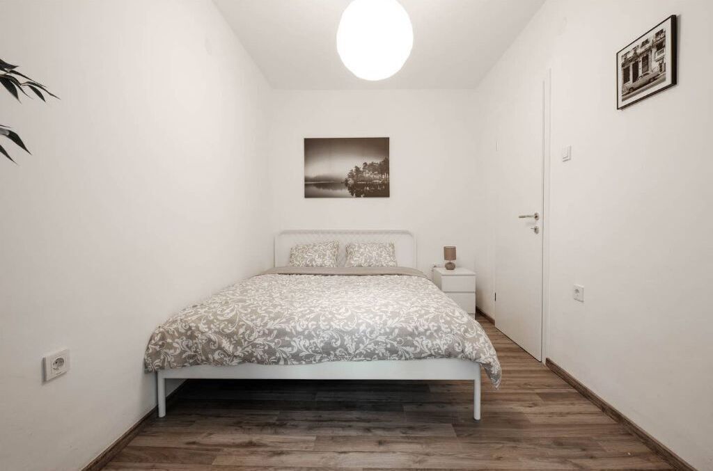 A bedroom with a bed, a bedside table, a nightstand, a wall-mounted picture, and a door.