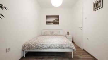 A bedroom with a bed, a bedside table, a nightstand, a wall-mounted picture, and a door.