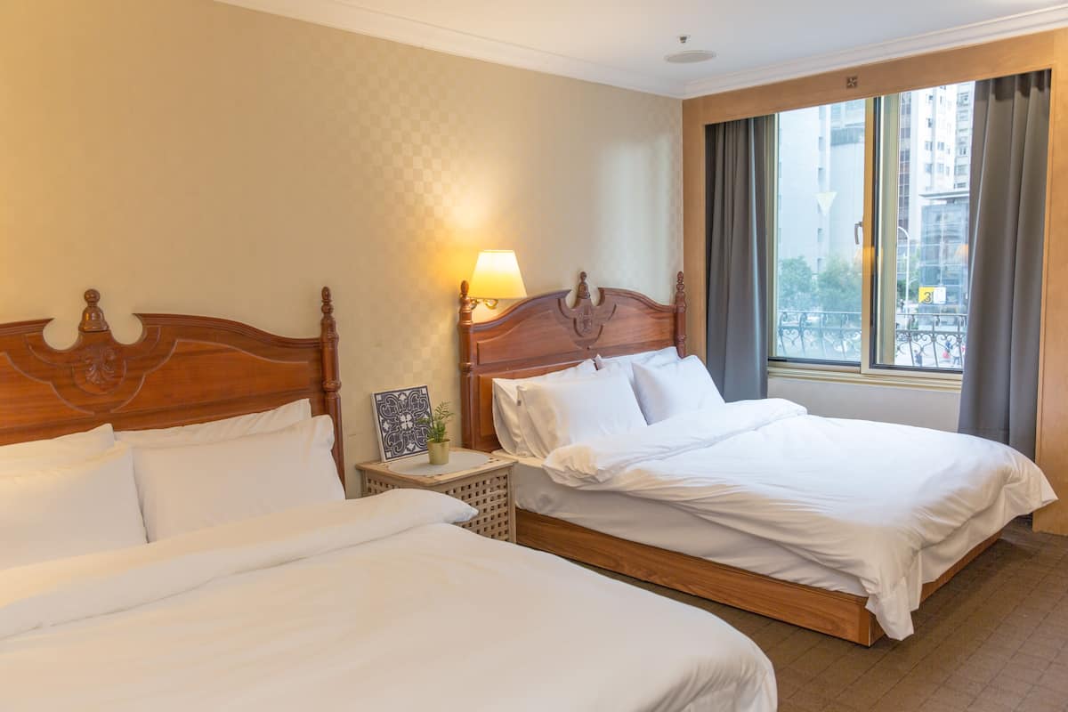 A hotel room with two beds, a nightstand with a lamp, a small plant, and a window with a view of buildings.