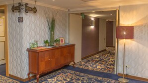 Un hall d’hôtel avec un papier peint à motifs, une console en bois agrémentée d’objets décoratifs, une moquette bleue et un lampadaire.