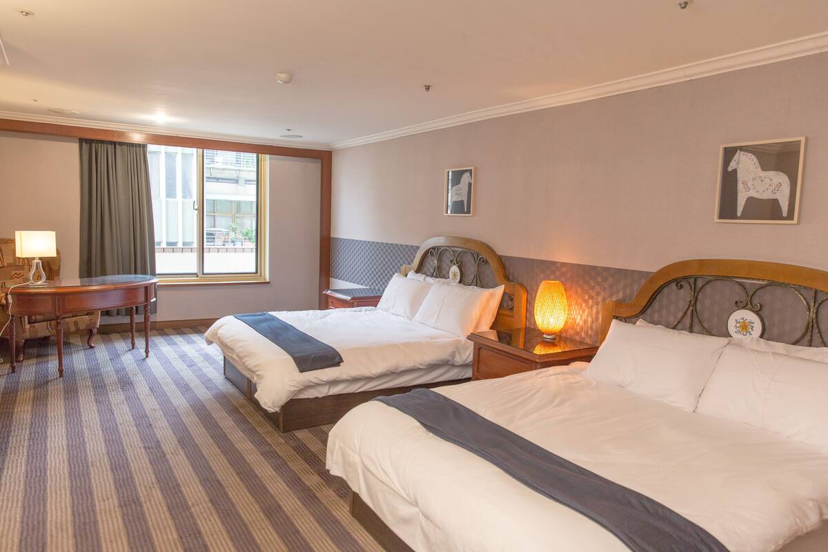 A hotel room with two beds, a desk, a lamp, and a window with curtains.