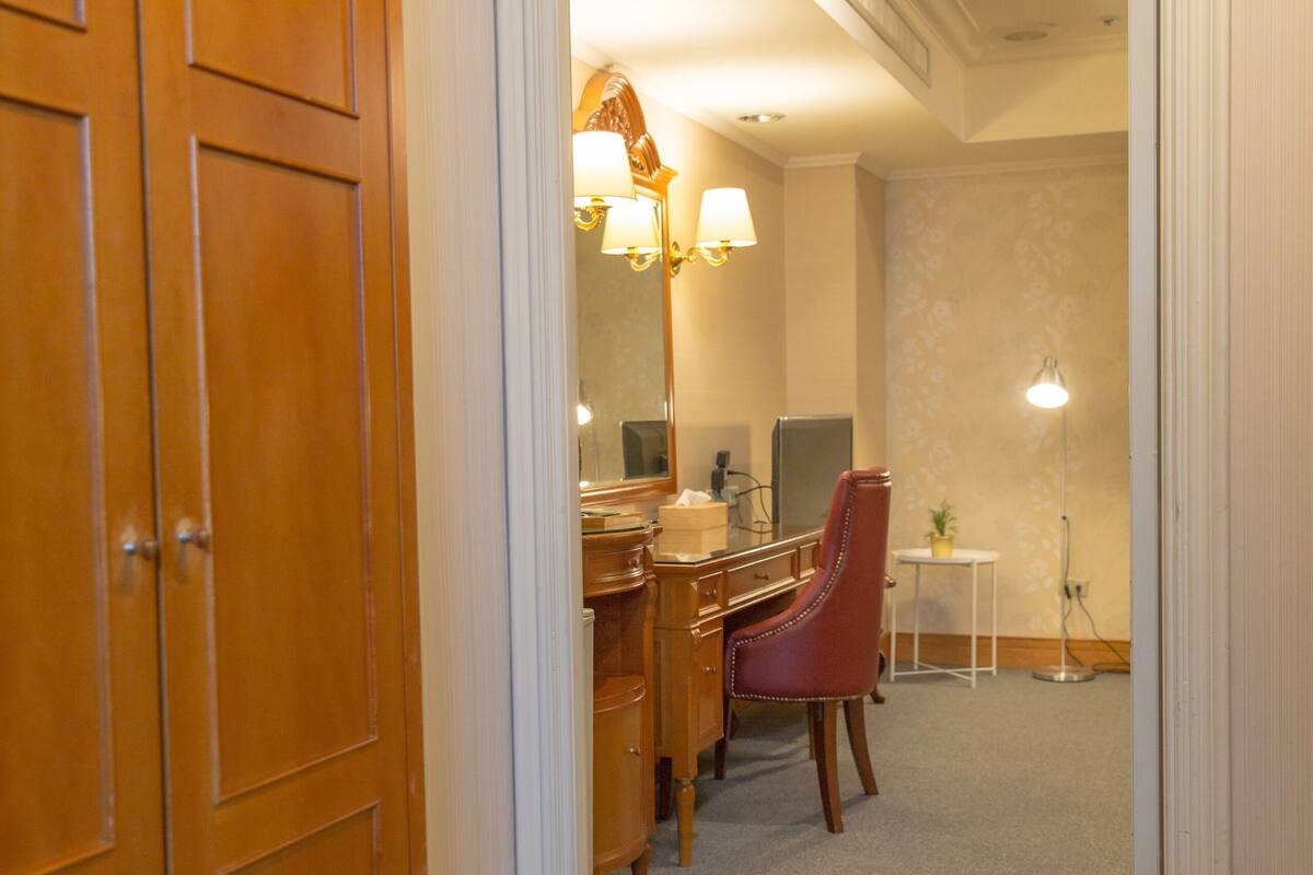 A hotel room with a wooden wardrobe, a desk with a chair, a small table with a lamp, and a mirror.