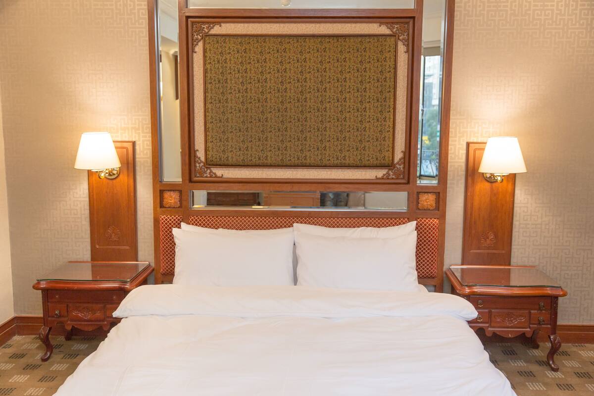 A hotel room with a large bed, two bedside tables with lamps, a wooden headboard with a mirror, and patterned wallpaper.