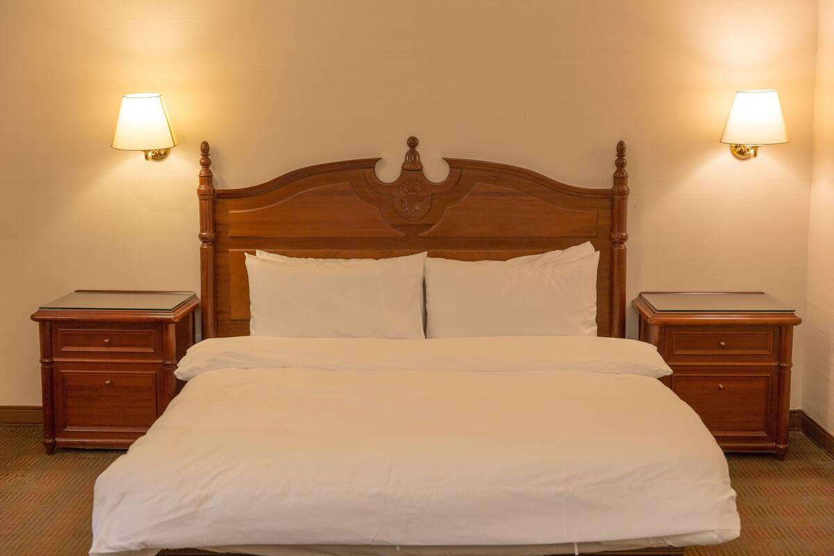 A neatly made bed with white linens, flanked by two wooden nightstands with lamps, in a room with beige walls.