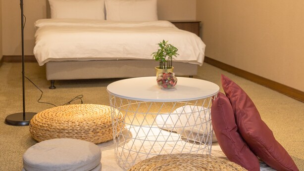 Une chambre à coucher avec un lit, une table de chevet avec une lampe, une petite plante et deux poufs en osier posés sur un tapis.