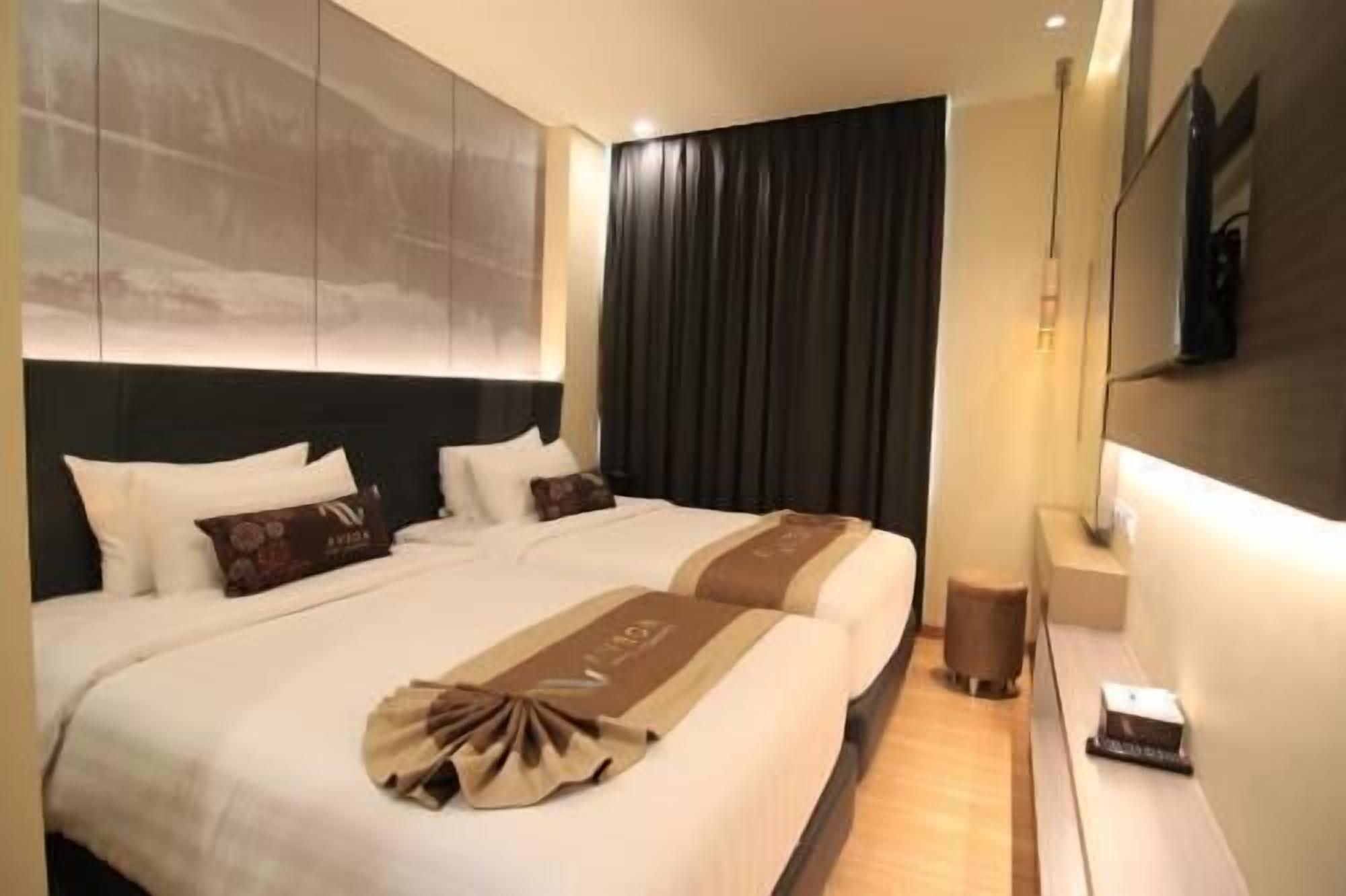 Ein Hotelzimmer mit einem großen Bett, dunklen Vorhängen, einem an der Wand montierten Fernseher und einer kleinen Mülltonne.