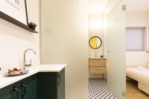 A narrow hallway with a sink and mirror on the left, a small bathroom with a toilet and sink on the right, and a bed with towels on the far right. - AirTLV - Levinski 113 Hotel - Hostel (Tel Aviv)