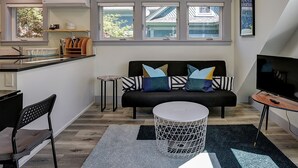 A compact living space with a kitchenette, a sofa with cushions, a TV, and a small round table. - Hawthorne Urban Loft Private ADU with Parking & AC (Portland)