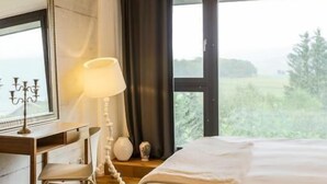 A hotel room with a large bed, a desk with a chair, a lamp, and a view of greenery through the window. - Hotel de l'Ours Preles (Plateau de Diesse)
