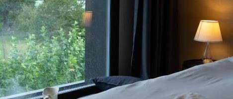 Habitación de hotel con una cama, una mesita de noche con lámpara, una ventana con vistas a zonas verdes y un edificio, y una taza sobre la mesa.