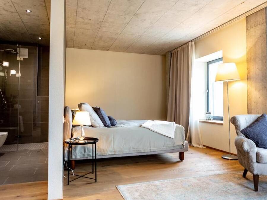 A modern bedroom with a bed, bedside table, chair, and a glass-enclosed shower area.