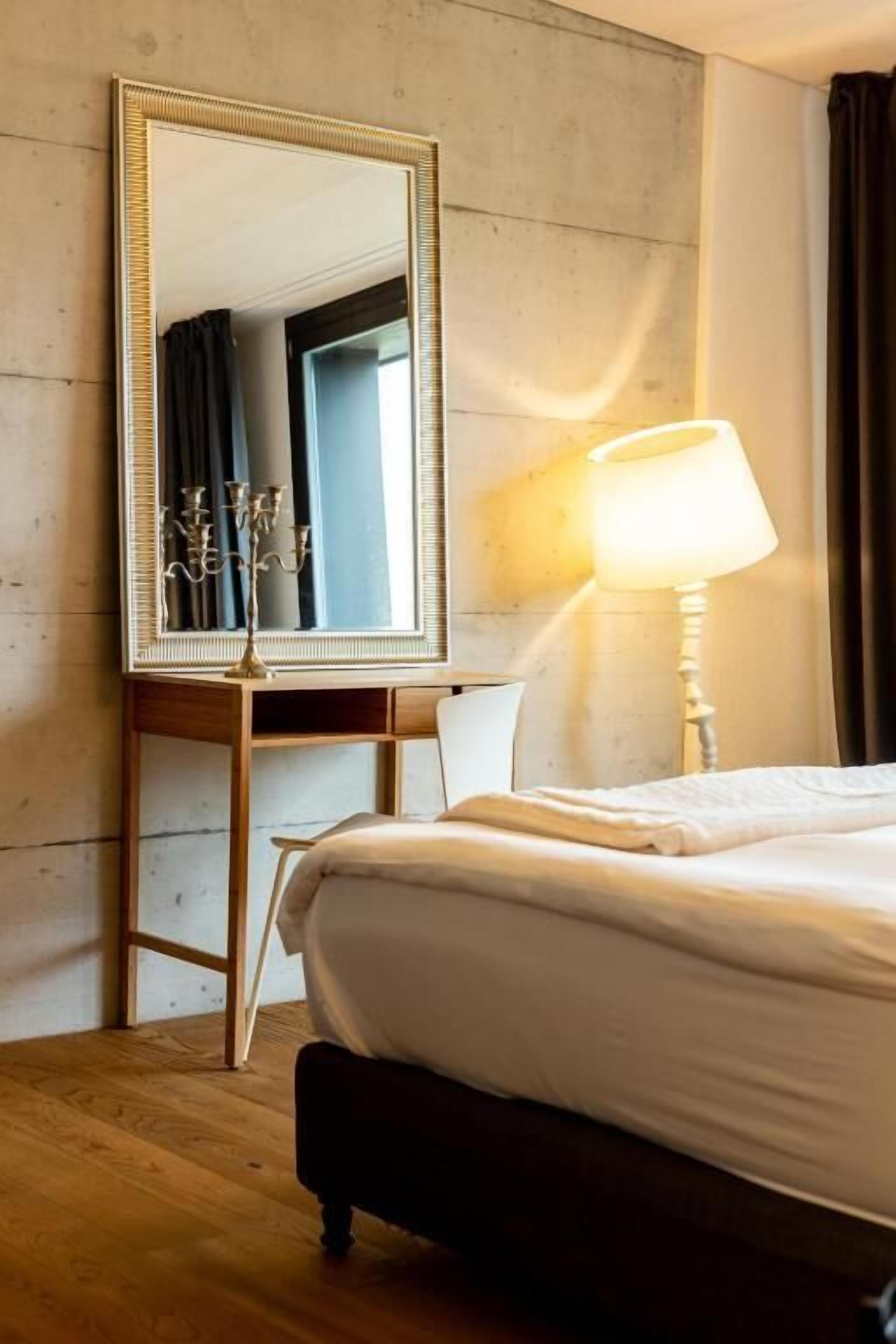 Una habitación de hotel con un espejo grande, una lámpara de noche, una cama y suelo de madera.