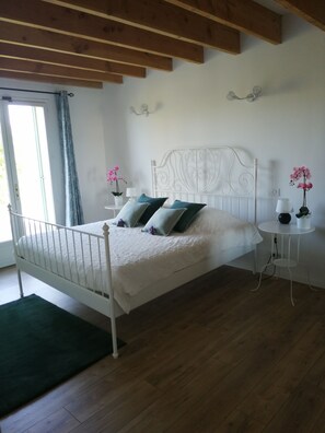 A bedroom with a white metal bed frame, a green rug, wooden flooring, a white bedside table with a lamp, and a window with curtains. - House with large garden, swimming pool, exceptional view and 300m from the town center (Vaison-la-Romaine)