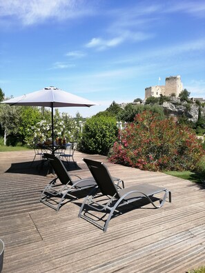 En træterrasse med liggestole og et bord under en hvid parasol, med udsigt til et slot og frodig vegetation.
