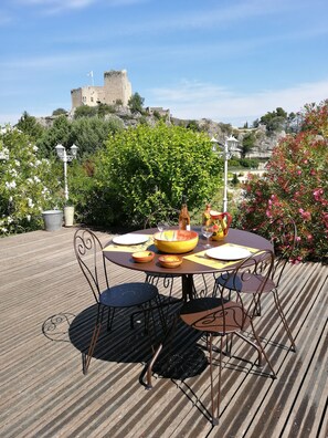 A round table with chairs on a wooden deck, a large bowl, and a pitcher, with a castle in the background. - House with large garden, swimming pool, exceptional view and 300m from the town center (Vaison-la-Romaine)