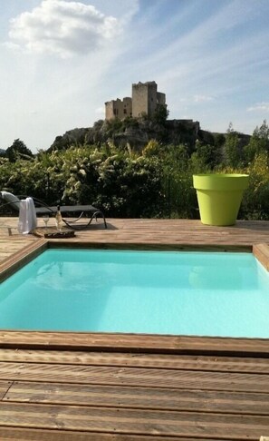 A swimming pool with a wooden deck, a lounge chair, and a large planter. In the background, there is a castle-like structure on a hill. - House with large garden, swimming pool, exceptional view and 300m from the town center (Vaison-la-Romaine)