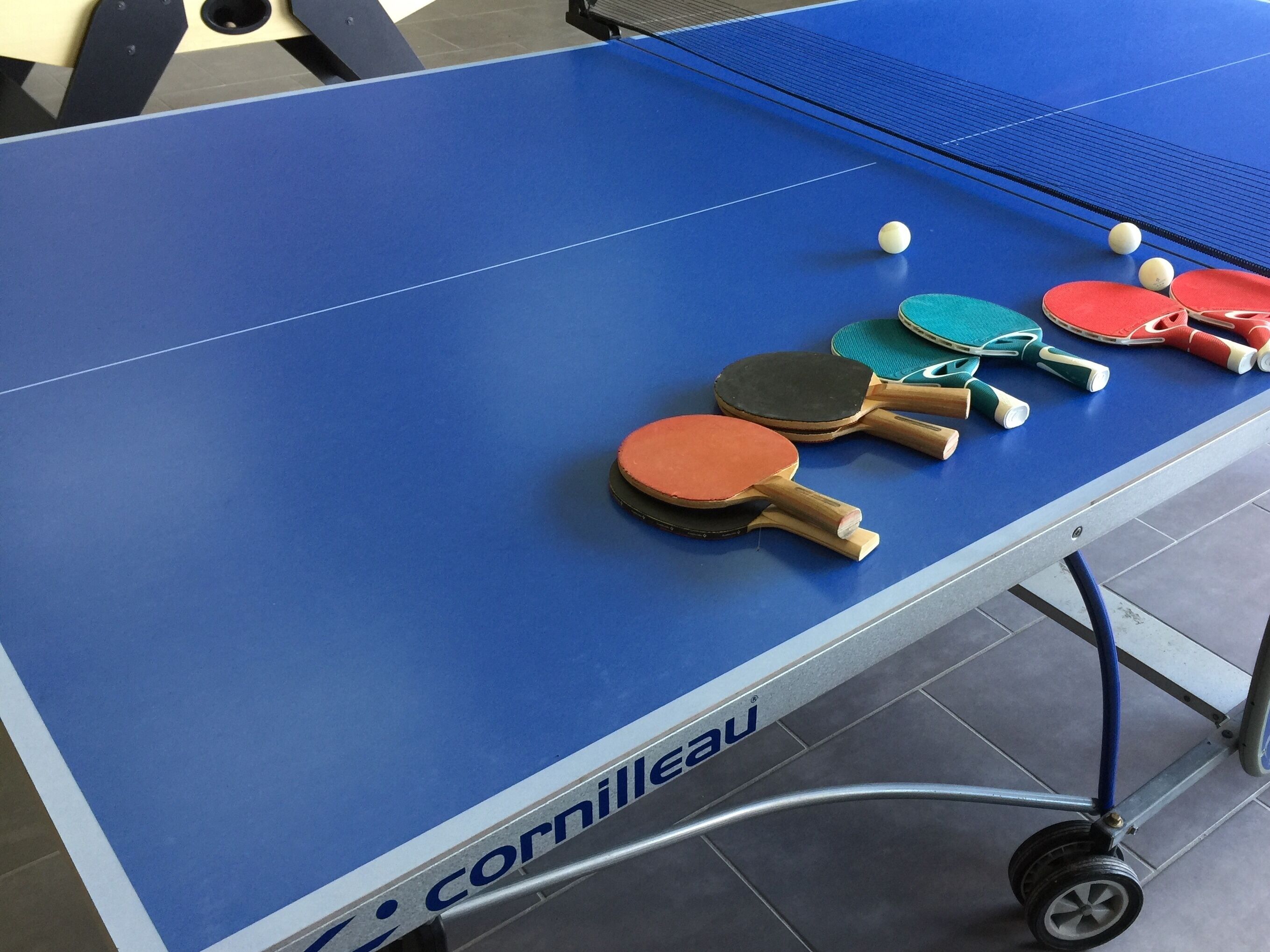 Une table de ping-pong dotée de plusieurs raquettes et balles, portant la marque « Cornilleau ».